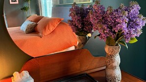 A room with a round mirror, a bed with orange bedding, a vase with purple flowers, a framed picture on the wall, and a small table with a box and a black rectangular object.