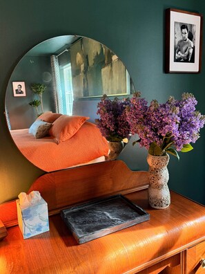 A room with a round mirror, a bed with orange bedding, a vase with purple flowers, a framed picture on the wall, and a small table with a box and a black rectangular object. - Brando King Room at Mountain Daddies B & B (Oakhurst)