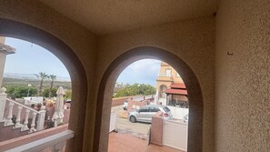Vista attraverso una finestra ad arco con un balcone, su una strada con auto parcheggiate e un edificio con tetto rosso.