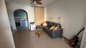 A living room with a sofa, a glass coffee table, a ceiling fan, a guitar, and a kitchen in the background.