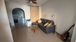 Un soggiorno con un divano, un tavolino in vetro, una ventola a soffitto, una chitarra e una cucina sullo sfondo.