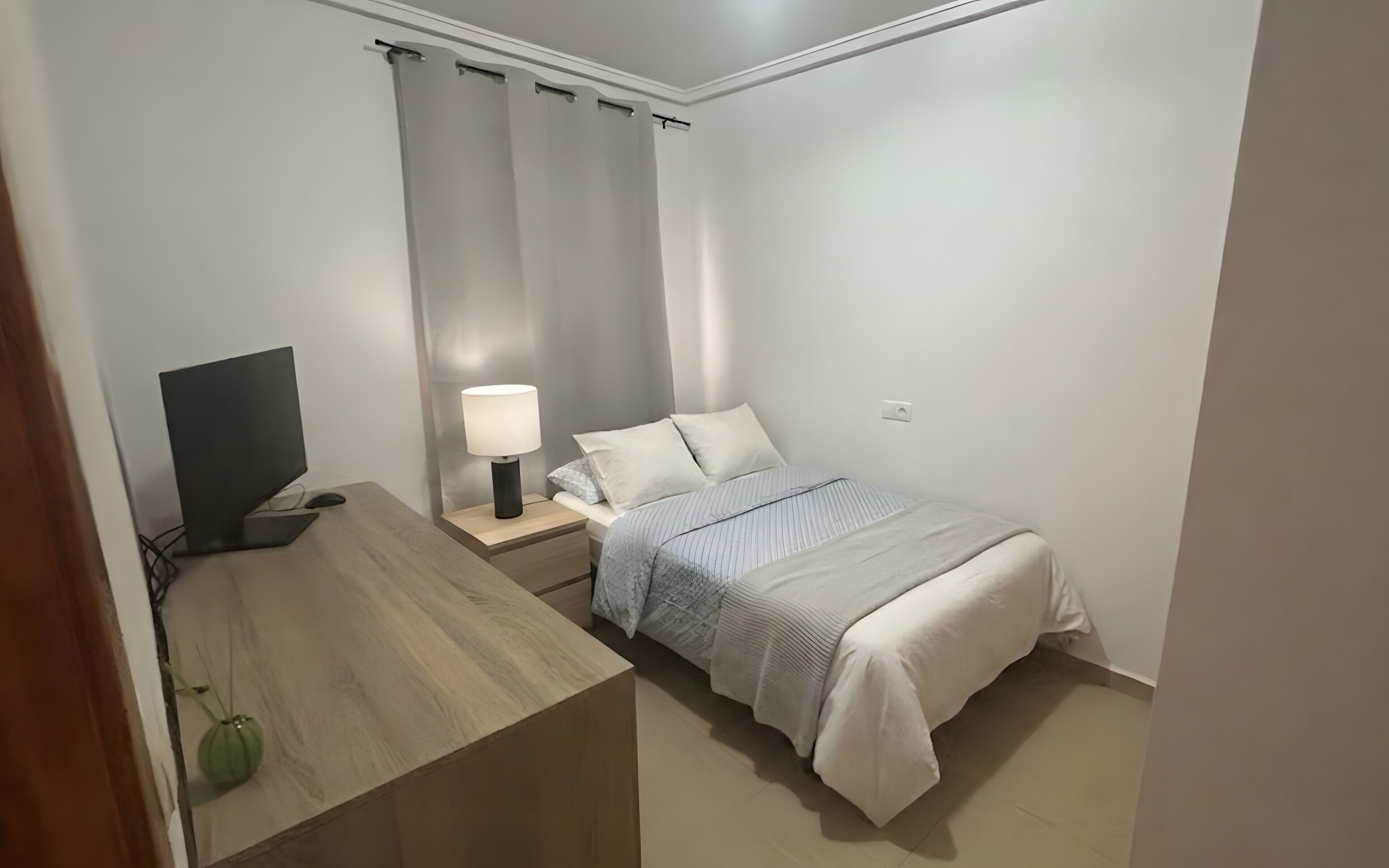 Un dormitori petit i ordenat amb llit, taula de nit, làmpada i televisió.