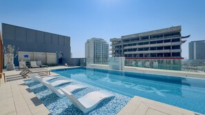 En pool på taget med liggestole, et overdækket opholdsområde og moderne bygninger i baggrunden.