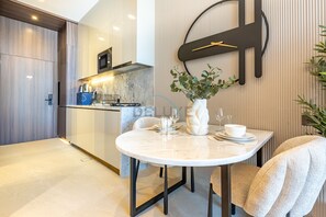 A modern kitchen with a built-in microwave, a stove, and a sink. There is a dining area with a round table and chairs, and a potted plant on the wall. - Stylish Modern Studio with Community Views, Next to the Community Park (Dubai)