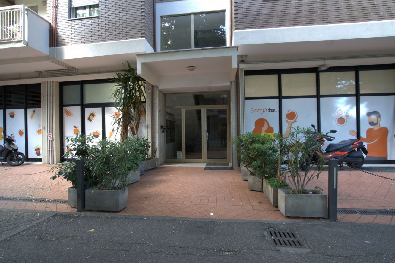 Un bâtiment avec une entrée en verre, des plantes en pot et un scooter garé à l’extérieur.