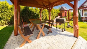 Un patio al aire libre con mesas y bancos de madera, una parrilla y una estructura de juegos al fondo.