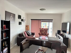 A living room with a sofa, armchair, dining table, bookshelf, and a TV. - Fourways Retreat Your Home Away From Home (Johannesburg)