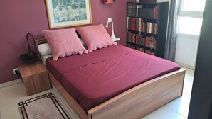 A bed with a wooden frame, a red bedspread, and two pink pillows. There is a white bedside table with a lamp, a bookshelf filled with books, and a framed picture on the wall. - Beautiful apartment with a piano (Sainte-Maxime)