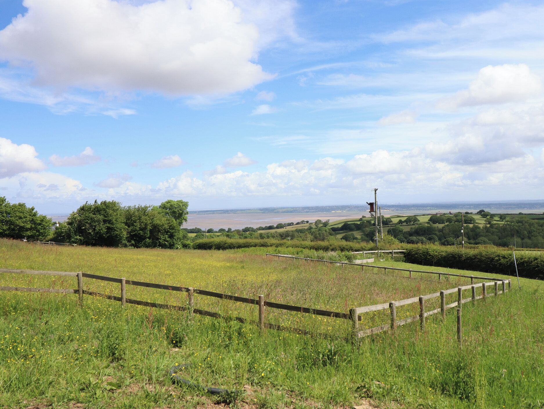 A rural landscape with a wooden fence, greenery, and a distant view of a body of water.
