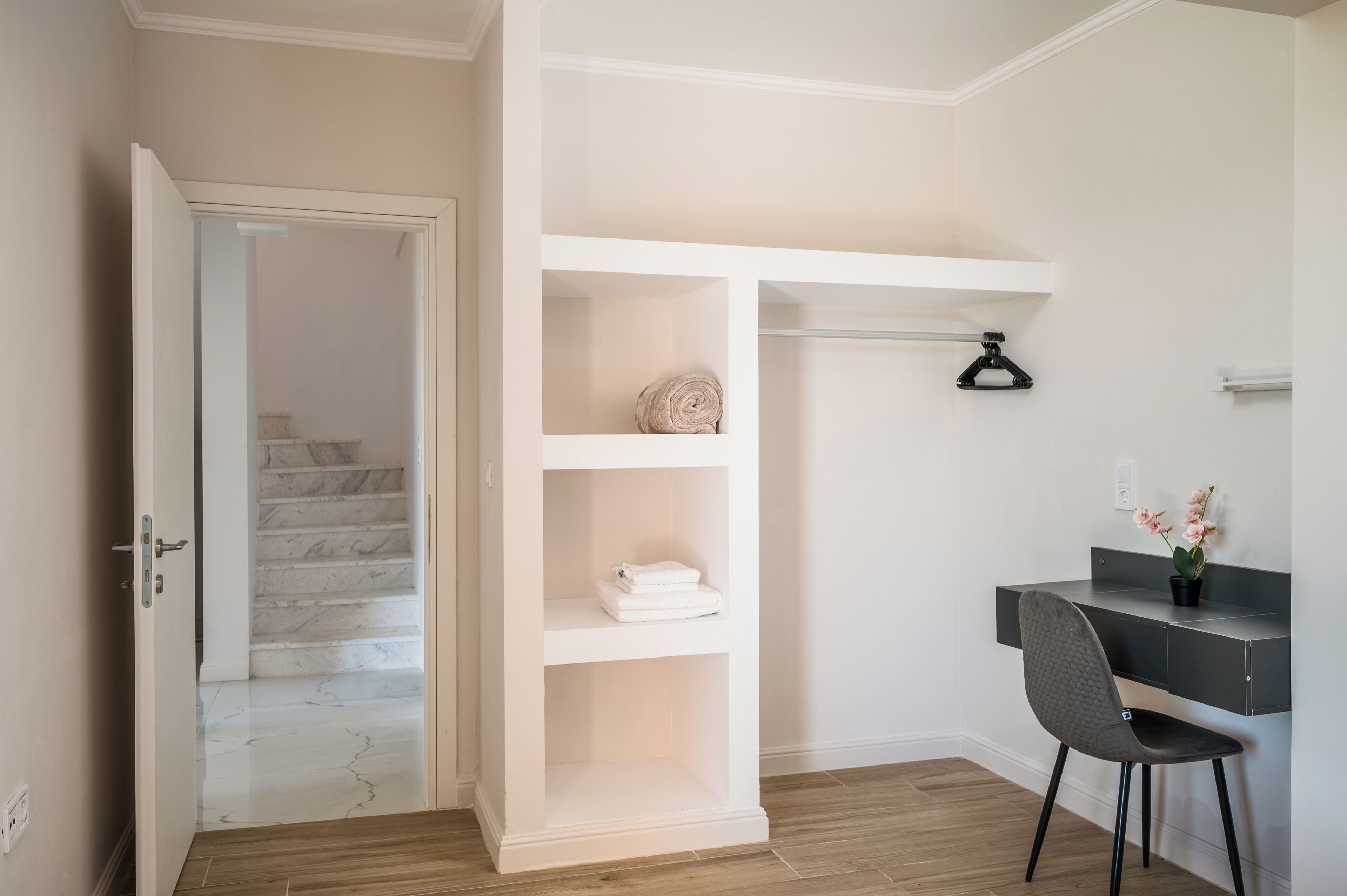 Ein Zimmer mit einer weißen, fest eingebauten Regaleinheit, einem Schreibtisch mit Stuhl, einer Topfpflanze und einer Treppe, die in ein weiteres Zimmer führt.