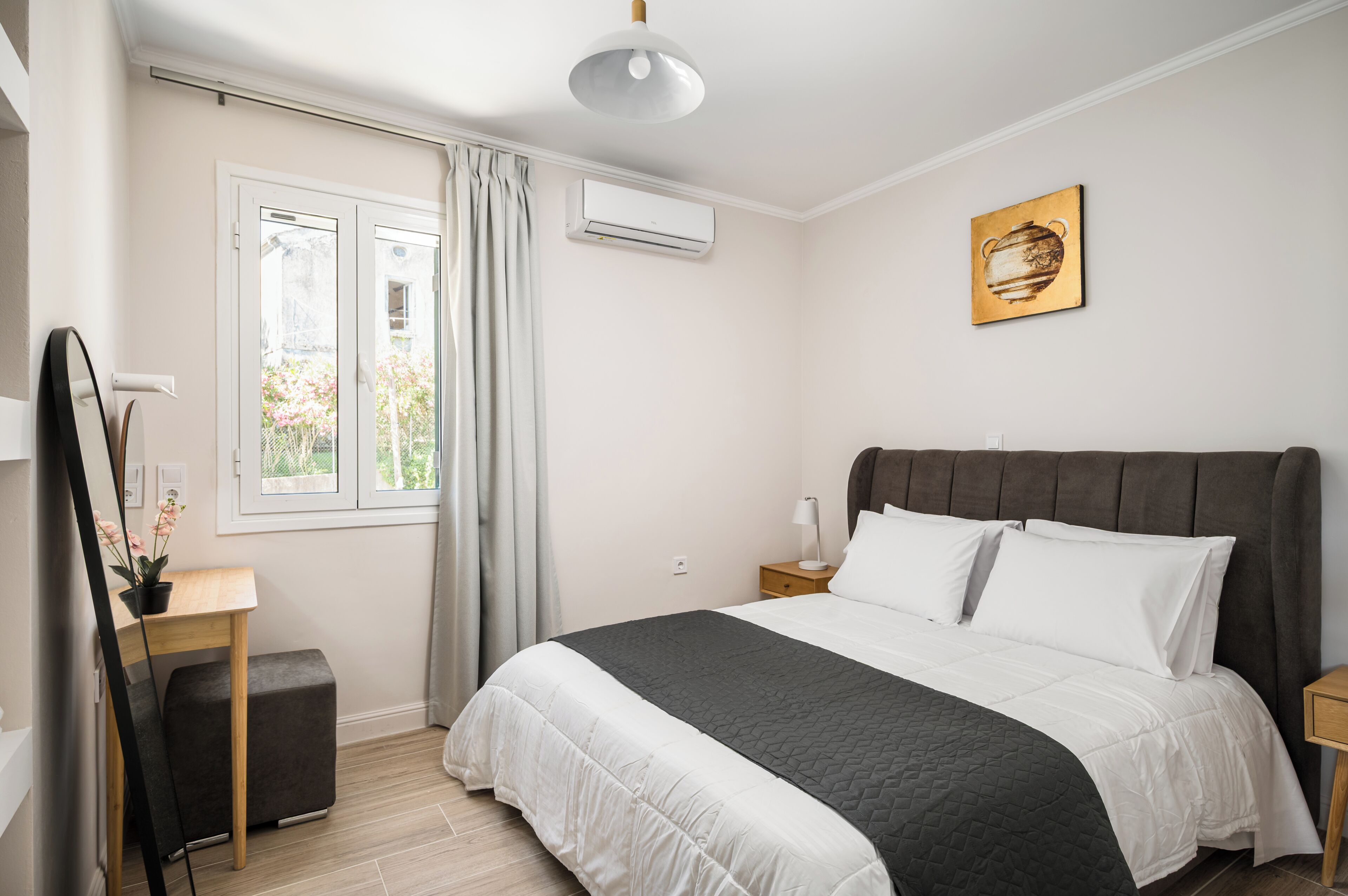 A bedroom with a bed, bedside tables, a desk, a mirror, and an air conditioning unit.