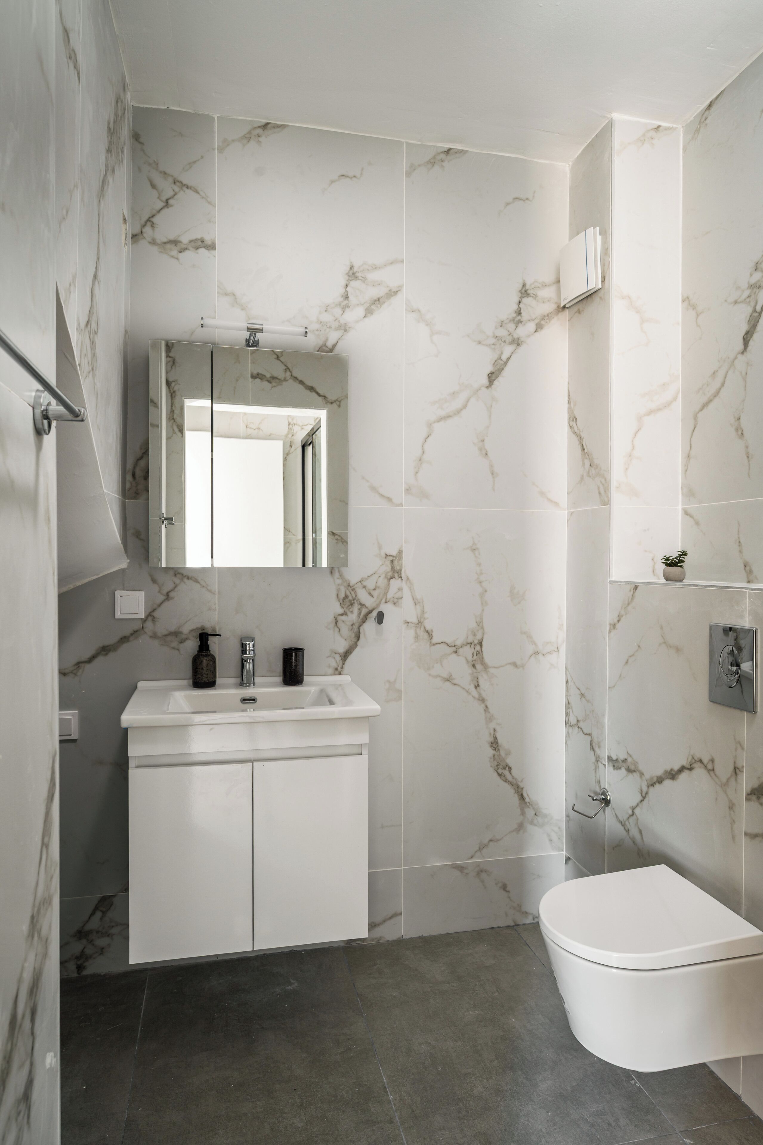Un baño con pared de mármol, un lavamanos blanco, un espejo y un inodoro.