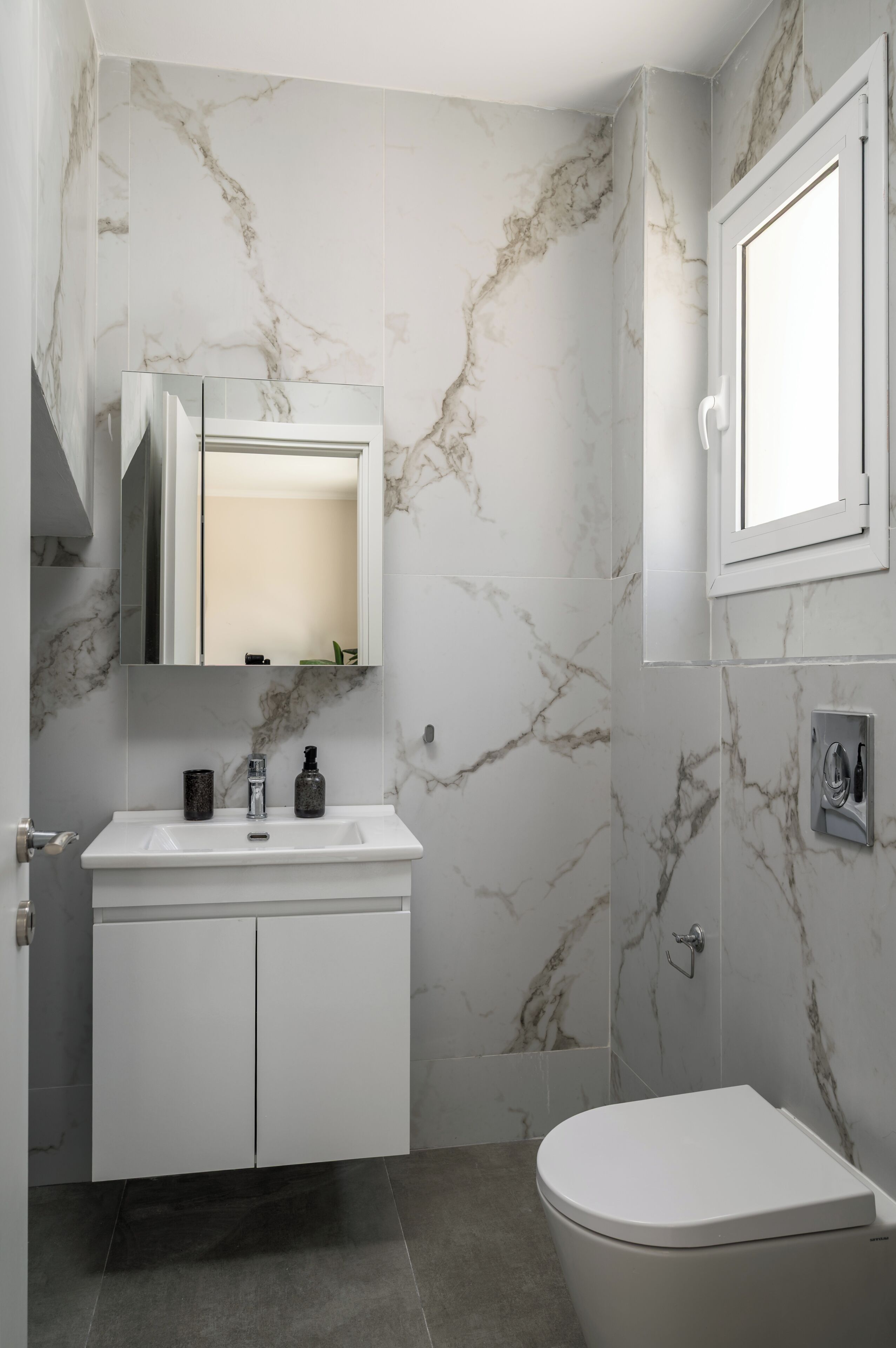 A bathroom with a marble wall, a white sink, a mirror, a toilet, and a window.