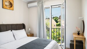 A bedroom with a bed, a nightstand, a bedside table with a lamp, a painting on the wall, and a balcony with a view of greenery and buildings.