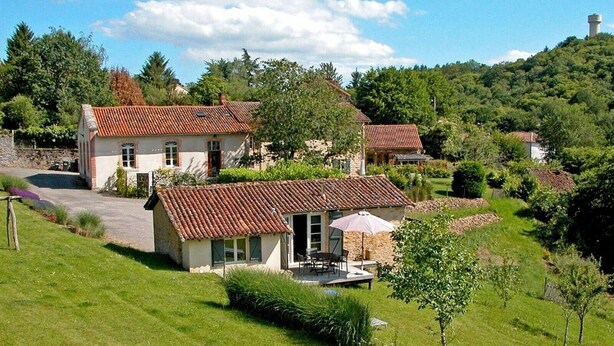 Une maison de campagne avec un toit en tuiles, une allée pavée et une petite terrasse aménagée avec du mobilier d’extérieur.