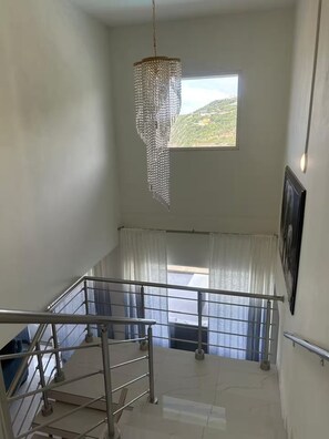 A staircase with metal railings, a hanging chandelier, a window with curtains, and a framed picture on the wall. - Belair Pool Villa w/ Rooftop & Island Views Oasis (Little Bay)