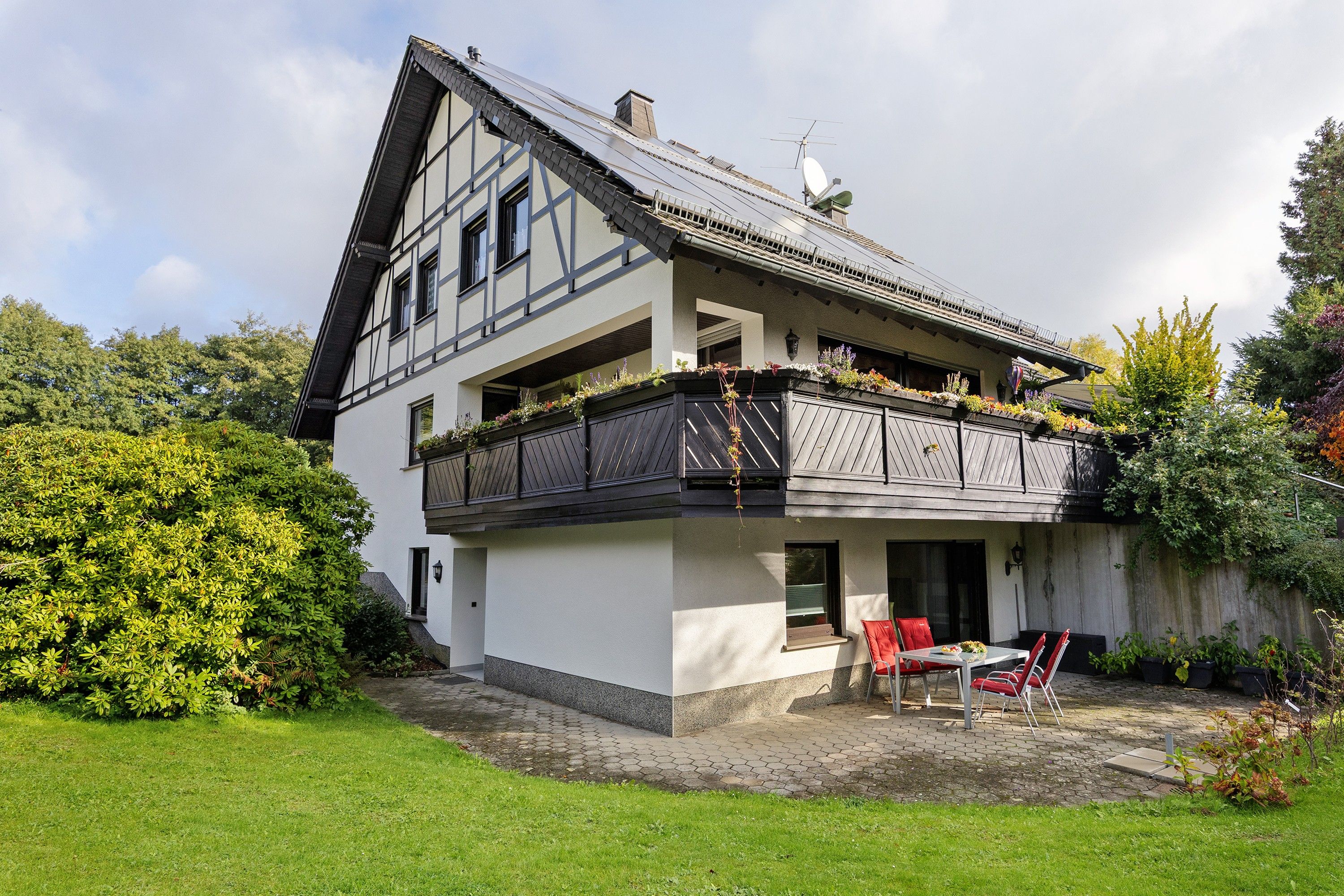 A two-story house with a balcony, a garden, and a patio with outdoor furniture.