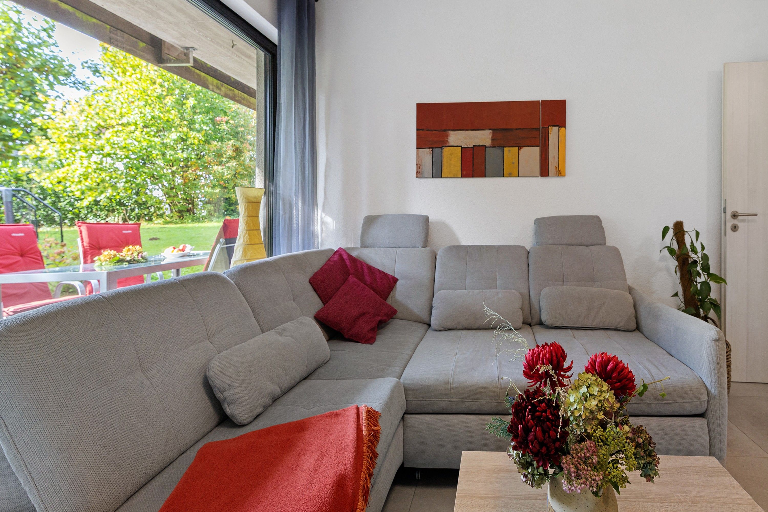 A modern living room with a grey sofa, red cushions, and a vase of flowers on a wooden table.