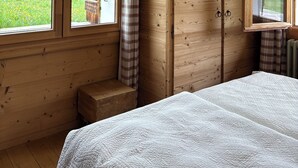 A wooden room with a bed, a window with a view of greenery, a plaid curtain, and a hanging lamp. - Charming Apartment for Rent - Log Cabin - Directly on Lake des Confins - Sleeps 8 (La Clusaz)