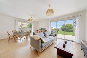 Un salón amplio con sofá gris, sillas de madera, una mesa de comedor y un ventanal con vistas al exterior.