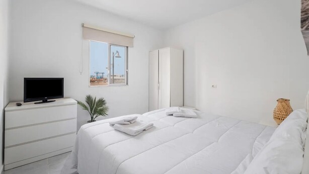 Un dormitorio con cama, televisor sobre una cómoda, ventana con vista a una obra en construcción y armario.