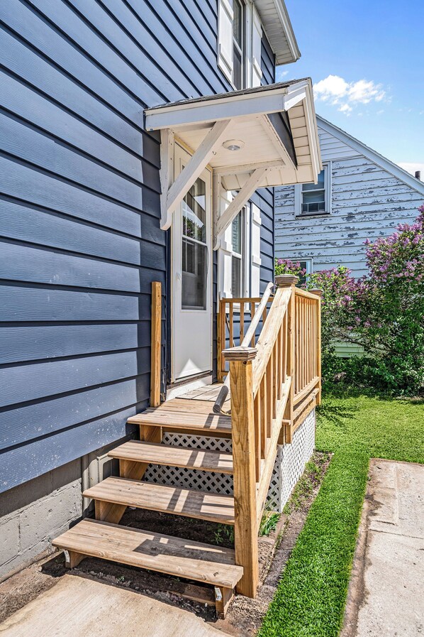 A wooden staircase leading to a house's entrance, a white door, and a small garden area. - Tour MQT, NMU - Comfy UP Getaway - Full Amenities - 1 Bed (Marquette)