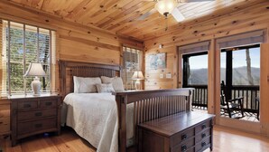 Un dormitorio de cabaña de madera con una cama, mesitas de noche, un banco y un balcón con vistas.