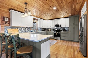 A modern kitchen with wooden ceiling, white cabinets, stainless steel appliances, and a central island with bar stools. - Blue Ridge Escape with Stunning Views, Fenced Yard & EV Charger (Blue Ridge)