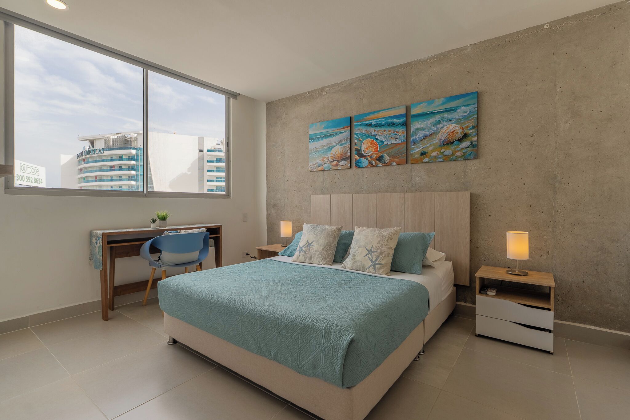 A modern bedroom with a large bed, a desk, and a view of buildings through a window.