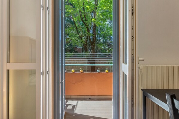 An open door leading to a balcony with a view of trees and a railing with potted plants.