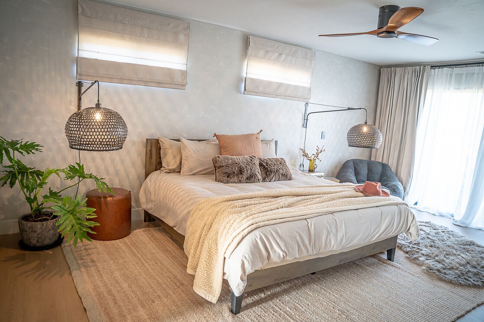 Une chambre à coucher avec un lit, des lampes de chevet, un ventilateur de plafond, une plante en pot et une fenêtre avec des rideaux.