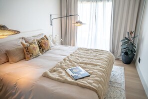 Ein ordentlich bezogenes Bett mit einem Buch, einer Lampe und einem Blumentopf in einem Zimmer mit einem großen Fenster und Vorhängen.