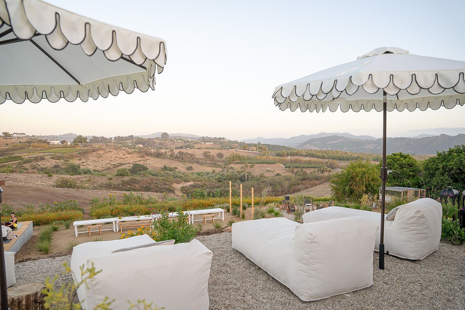 Un área de estar al aire libre con sillas y mesas blancas acolchadas, sombrillas blancas y una vista panorámica de colinas y montañas.