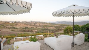 Ein Sitzbereich im Freien mit weißen, gepolsterten Stühlen und Tischen, weißen Sonnenschirmen und einer malerischen Aussicht auf sanfte Hügel und Berge.