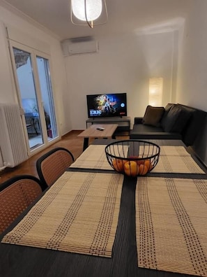 Ein Wohnzimmer mit Fernseher, einer Couch, einem Couchtisch mit Obstschale und einer Glastür zum Außenbereich.
