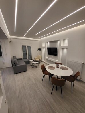 Ein modernes Wohnzimmer mit einer Couch, einem runden Esstisch mit Stühlen, einem an der Wand montierten Fernseher und einer Stehlampe.