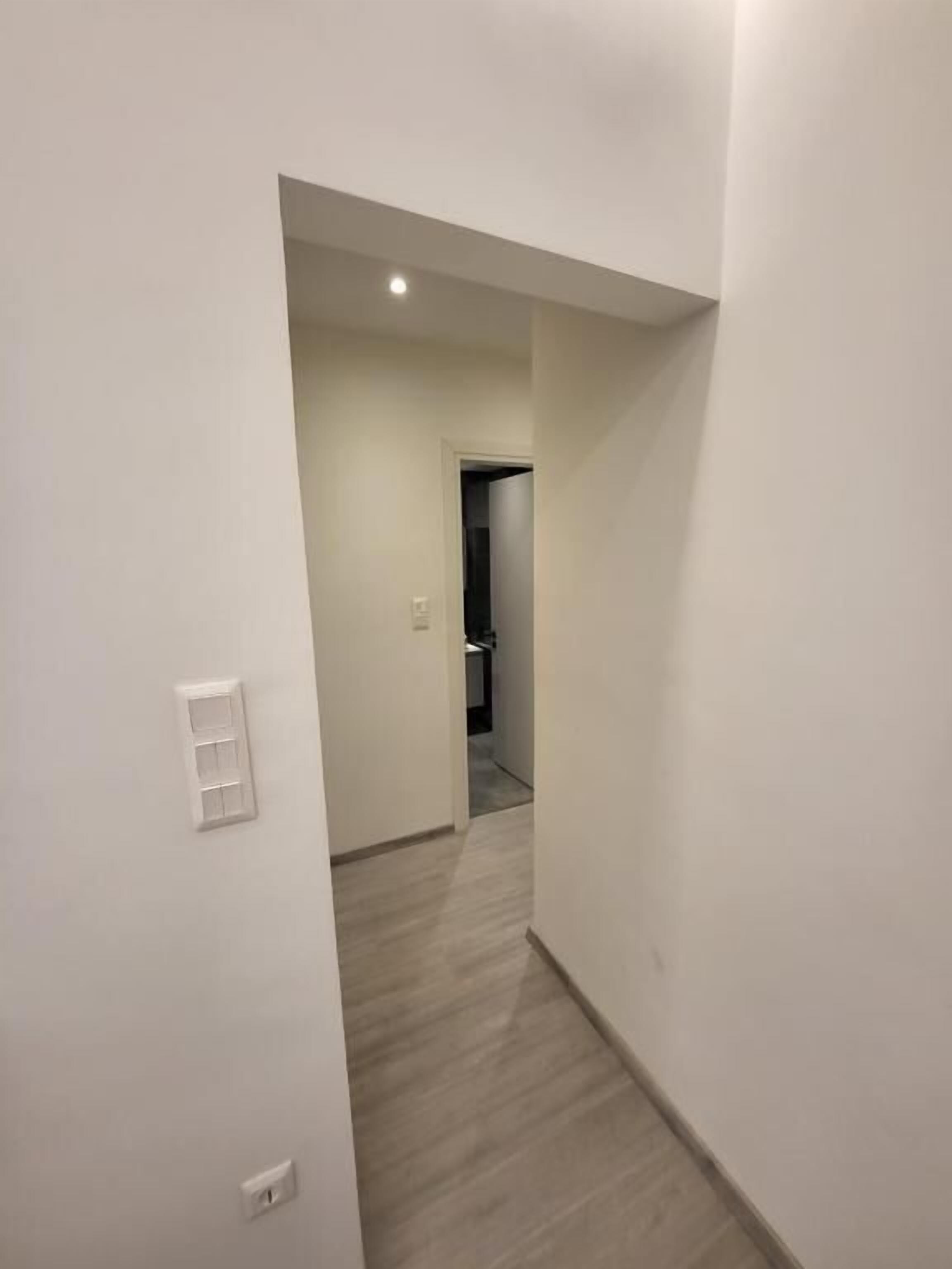 Un corridoio stretto con una porta in fondo, un interruttore sulla parete e un lampadario al soffitto.