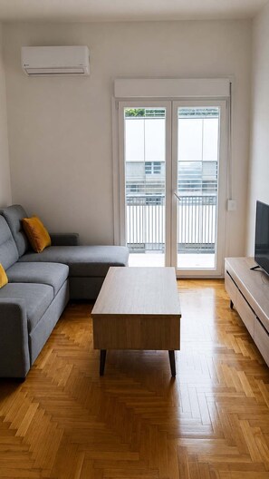 A living room with a grey sofa, a wooden coffee table, a TV on a stand, and a sliding glass door leading to a balcony. - Boutique 1 Bd Stay in Mavili Square Athens (Athina)