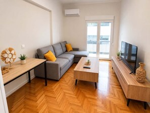 A modern living room with a grey sofa, wooden coffee table, TV stand, and a large window with blinds. - Boutique 1 Bd Stay in Mavili Square Athens (Athina)