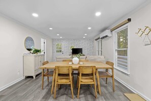 A dining area with a wooden table and chairs, a white brick wall, a TV, and a window with blinds.