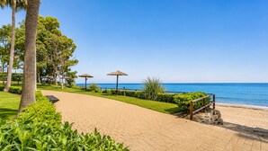 Ein gepflasterter Weg führt zu einem Strand mit Palmen und Reetsonnen.