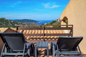 Eine Terrasse mit zwei Liegestühlen und einem Tisch, mit Blick auf eine Küstenlandschaft mit Gebäuden und Hügeln.