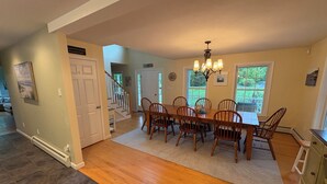 A dining area with a wooden table and chairs, a chandelier, and a view of the outdoors through large windows. - New- Spacious 4-bedroom Winter Rental Home with Fireplace in Wells, Maine (Wells)