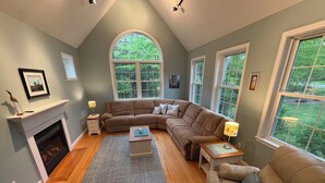 A living room with a fireplace, a sectional sofa, a coffee table, and large windows. - New- Spacious 4-bedroom Winter Rental Home with Fireplace in Wells, Maine (Wells)
