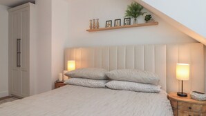 A bedroom with a bed, bedside lamps, a nightstand, a wardrobe, and a shelf with decorative items. - Puffin Lodge (Filey)