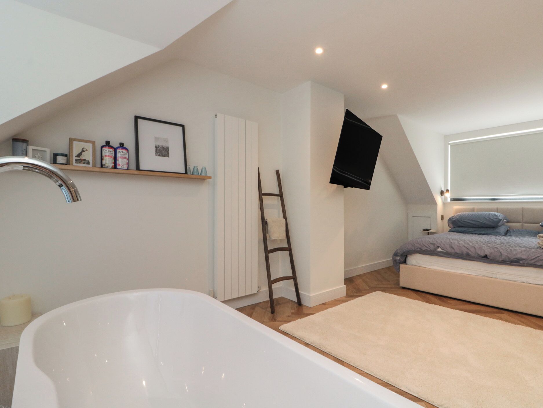 Un dormitorio con una bañera blanca, una cama con edredón gris, una escalera de madera y un estante con marcos y botellas.