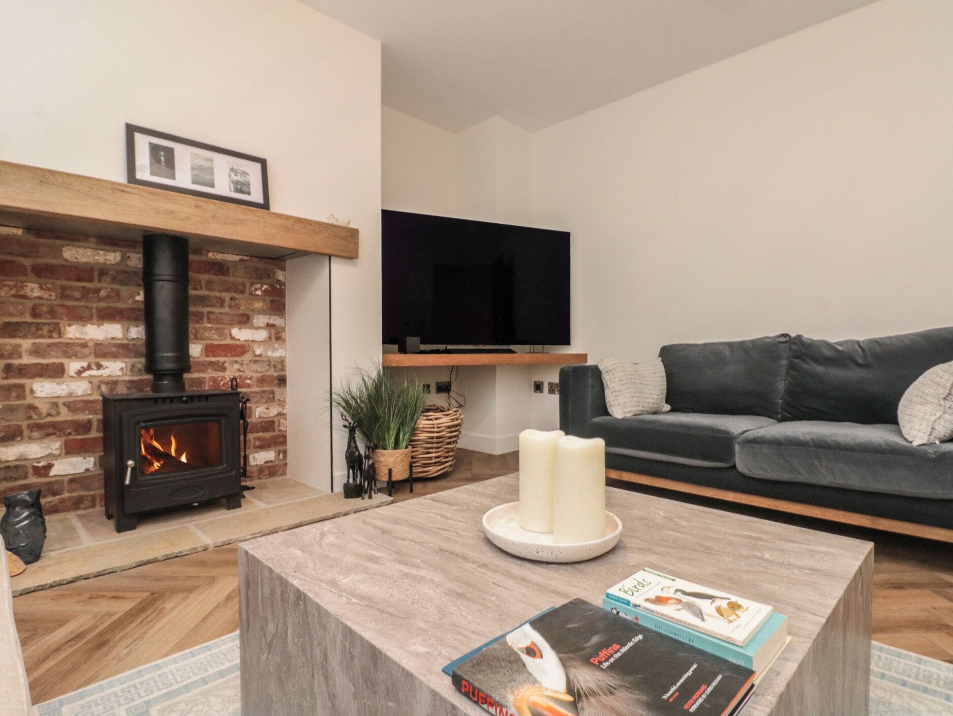 A living room with a brick fireplace, a flat-screen TV, a grey sofa, and a coffee table with books.