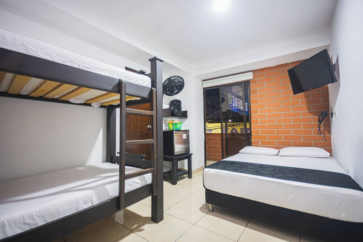 A dorm-style room with bunk beds, a desk with a computer, a TV mounted on the wall, and a brick-patterned wall.