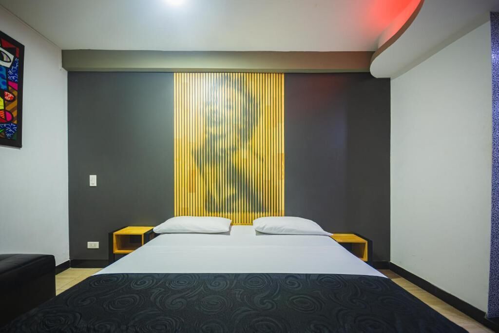 Habitación de hotel con cama, dos mesitas de noche, una pared con persianas verticales y una obra de arte colorida en la pared izquierda.