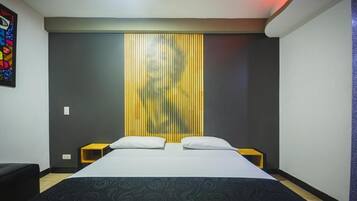Habitación de hotel con cama, dos mesitas de noche, una pared con persianas verticales y una obra de arte colorida en la pared izquierda.